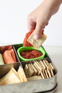 child's hand dipping crackers and pepperoni in pizza sauce for punchable in stainless steel container