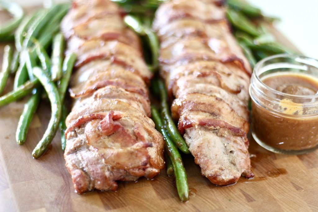 bacon wrapped pork tenderloin on cutting board with green beans and sauce