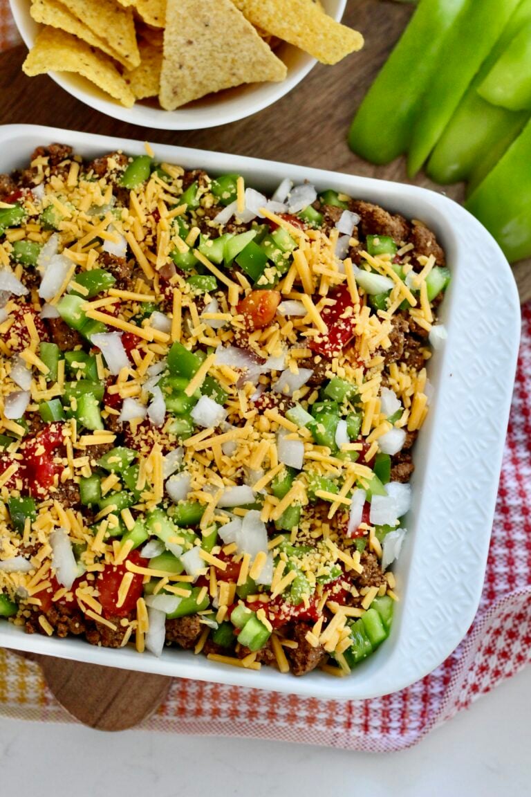 high protein taco dip 4 | Milk & Honey Nutrition White baking dish of layered high protein taco dip topped with diced onions, green peppers, tomatoes and shredded cheddar; tortilla chips nearby.