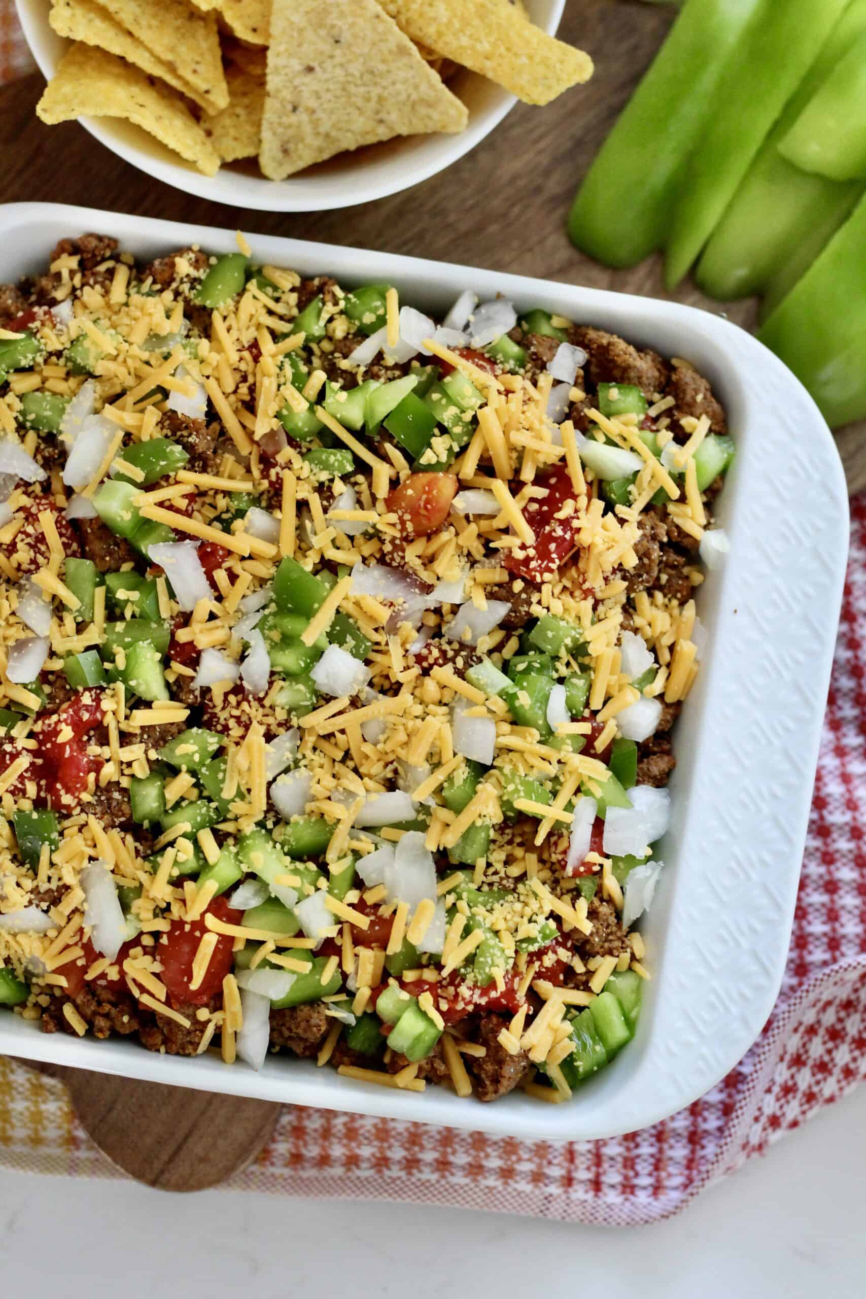 high protein taco dip 4 | Milk & Honey Nutrition White baking dish of layered high protein taco dip topped with diced onions, green peppers, tomatoes and shredded cheddar; tortilla chips nearby.