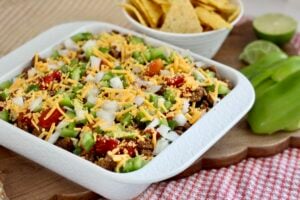 high protein taco dip 6 | Milk & Honey Nutrition Loaded high protein taco dip in a white baking dish topped with cheddar, onions, green peppers, and tomatoes; chips in a bowl behind.
